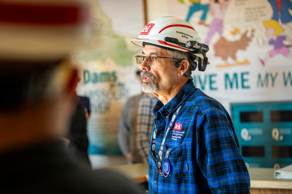 A man wearing a white US Army Corps of Engineers hard hat and blue plaid shirt is speaking or presenting in what appears to be a visitor center or educational facility. His hard hat reads 'Building Strong Safely' and his shirt patch identifies him as being from the 'Gavins Point Project.' He appears to be addressing a small group in a room with informational posters about dams visible in the background.