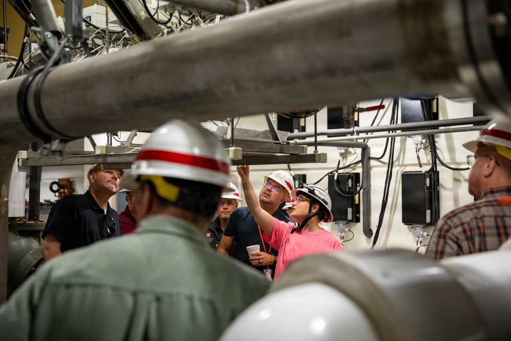 A group of people wearing hard hats are gathered inside an industrial facility, likely on a guided tour or safety briefing. A woman in a pink shirt is pointing upward toward overhead pipes and equipment, appearing to explain or demonstrate something to the group. The setting features extensive piping, cables, conduits, and mounted electronic equipment on the walls and ceiling.