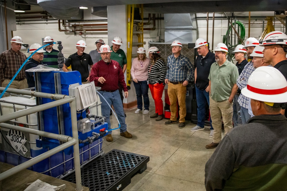 A group of approximately 20 people wearing white hard hats are gathered in an industrial facility for what appears to be a guided tour or safety briefing. A man in a red shirt is presenting or explaining equipment to the group. The setting appears to be a mechanical or utility room with pipes, industrial equipment, and a blue IBC tote container marked with hazmat placard number 3082.