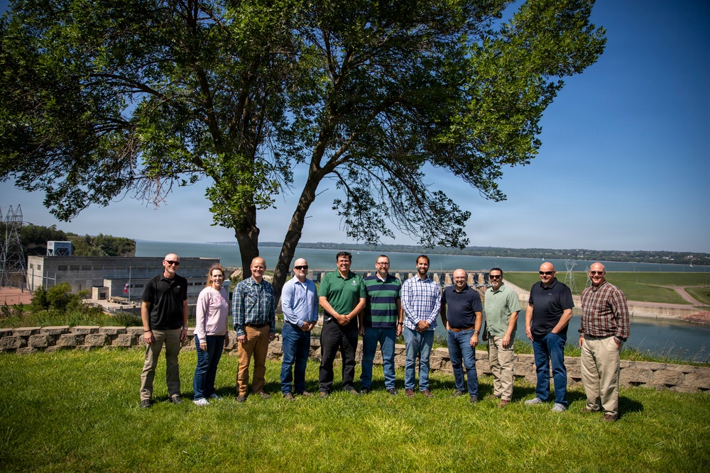 A group of approximately twelve adults poses for a formal group photograph on a grassy hillside overlooking a dam and reservoir. The setting appears to be an outdoor site visit or official gathering at a water infrastructure facility. The photo is taken on a clear, sunny day with a large tree providing partial backdrop.