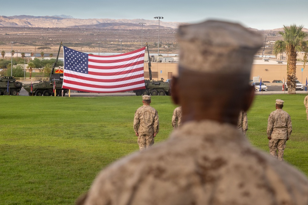 Military personnel in desert camouflage uniforms stand at attention on a grass parade field facing a large American flag displayed between two armored military vehicles. The setting appears to be a formal military ceremony at a desert installation, likely in the American Southwest based on the arid mountain terrain visible in the background. Palm trees, military equipment, and base infrastructure are visible in the background.