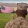 Military personnel in desert camouflage uniforms stand at attention on a grass parade field facing a large American flag displayed between two armored military vehicles. The setting appears to be a formal military ceremony at a desert installation, likely in the American Southwest based on the arid mountain terrain visible in the background. Palm trees, military equipment, and base infrastructure are visible in the background.