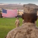 Military personnel in desert camouflage uniforms stand at attention on a grass parade field facing a large American flag displayed between two armored military vehicles. The setting appears to be a formal military ceremony at a desert installation, likely in the American Southwest based on the arid mountain terrain visible in the background. Palm trees, military equipment, and base infrastructure are visible in the background.