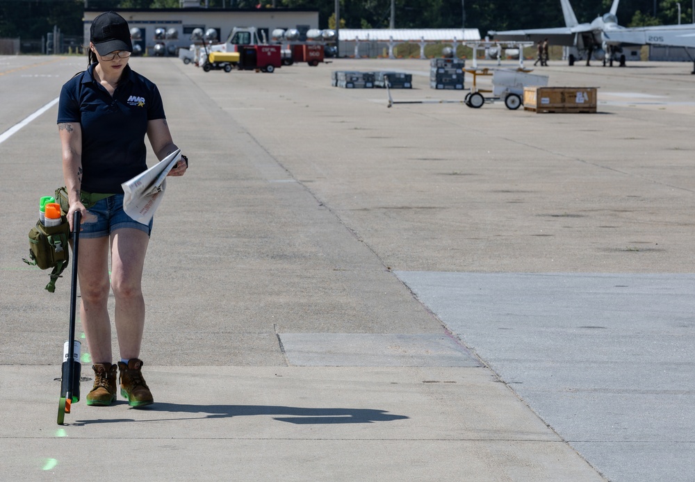 A woman wearing a navy blue polo shirt with 'NVR' or similar logo, denim shorts, and work boots is conducting a survey or inspection on a military airfield tarmac. She is holding a pole-mounted instrument (likely a ground-penetrating radar or measurement device) and reviewing documents simultaneously. Military aircraft and ground support equipment are visible in the background.