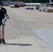 A woman wearing a navy blue polo shirt with 'NVR' or similar logo, denim shorts, and work boots is conducting a survey or inspection on a military airfield tarmac. She is holding a pole-mounted instrument (likely a ground-penetrating radar or measurement device) and reviewing documents simultaneously. Military aircraft and ground support equipment are visible in the background.