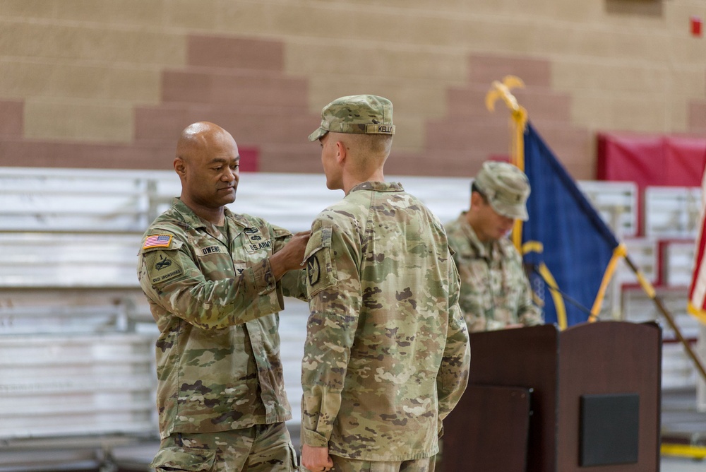 A U.S. Army ceremony taking place in what appears to be a gymnasium or indoor military facility. A senior soldier, identified by a name tape reading 'OWENS,' is pinning something onto the chest or collar of another soldier facing away from the camera, whose name tape reads 'KELLY.' A third soldier and a unit guidon flag are visible in the background.