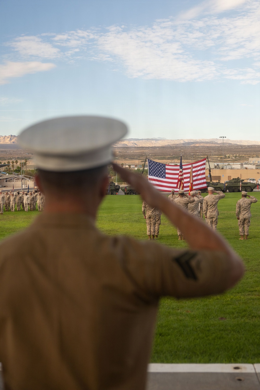 A U.S. Marine in the foreground, wearing a white dress cover and tan service uniform, renders a salute toward a large American flag displayed on a parade field. Multiple other Marines in camouflage uniforms are also saluting in formation on the grass. The setting appears to be a military base in a desert environment, with armored vehicles and mountains visible in the background.