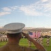 A U.S. Marine in the foreground, wearing a white dress cover and tan service uniform, renders a salute toward a large American flag displayed on a parade field. Multiple other Marines in camouflage uniforms are also saluting in formation on the grass. The setting appears to be a military base in a desert environment, with armored vehicles and mountains visible in the background.