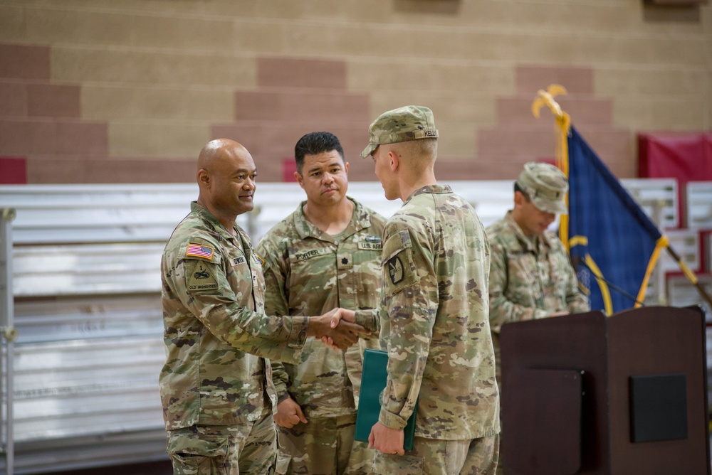 A military ceremony is taking place in what appears to be an indoor facility, likely a gymnasium or armory. A senior soldier on the left is shaking hands with another soldier on the right, who is holding a green folder, suggesting an award or recognition presentation. A third soldier wearing a U.S. Army patch and rank insignia stands between them as a witness or presiding officer.