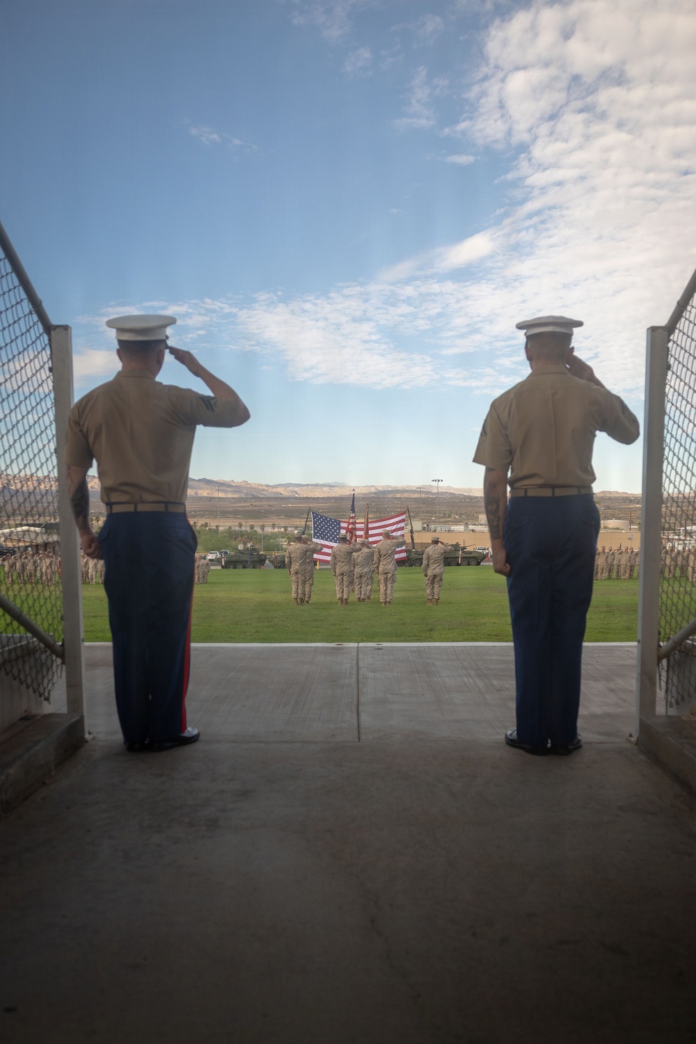 Two U.S. Marines in dress uniform stand at salute on an elevated platform, facing a formal military parade field where a color guard is presenting the American flag. The ceremony appears to be taking place at a desert military installation, with armored vehicles (likely LAV or Stryker variants) flanking the field and large formations of troops visible on both sides. The composition frames the two saluting Marines in the foreground against the expansive ceremony below.