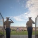 Two U.S. Marines in dress uniform stand at salute on an elevated platform, facing a formal military parade field where a color guard is presenting the American flag. The ceremony appears to be taking place at a desert military installation, with armored vehicles (likely LAV or Stryker variants) flanking the field and large formations of troops visible on both sides. The composition frames the two saluting Marines in the foreground against the expansive ceremony below.