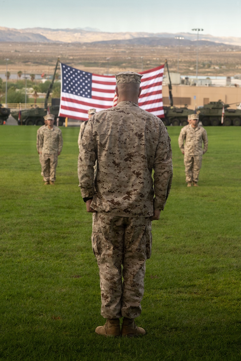 A U.S. Marine in desert MARPAT camouflage uniform stands at attention on a grass parade field, facing a large American flag suspended between two cranes or lift vehicles. Several other Marines in similar uniforms are positioned in the background, suggesting a formal military ceremony. The setting appears to be a military installation in an arid, desert region with mountains visible in the background.