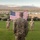 A U.S. Marine in desert MARPAT camouflage uniform stands at attention on a grass parade field, facing a large American flag suspended between two cranes or lift vehicles. Several other Marines in similar uniforms are positioned in the background, suggesting a formal military ceremony. The setting appears to be a military installation in an arid, desert region with mountains visible in the background.