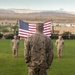 A U.S. Marine in desert MARPAT camouflage uniform stands at attention on a grass parade field, facing a large American flag suspended between two cranes or lift vehicles. Several other Marines in similar uniforms are positioned in the background, suggesting a formal military ceremony. The setting appears to be a military installation in an arid, desert region with mountains visible in the background.