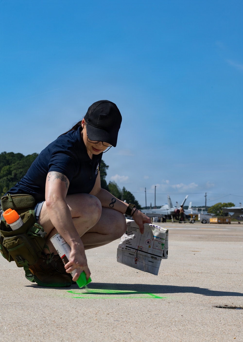 A woman in a dark navy shirt and black cap is crouching on a concrete airfield surface, using green spray paint to mark a shape on the ground. She is holding what appears to be a printed document or map in her other hand, and military aircraft are visible in the background. The setting appears to be a military air base or airfield.