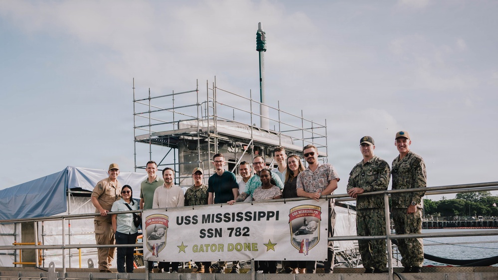 A group of approximately 15 people, including uniformed U.S. Navy personnel and civilians, pose for a photo in front of a banner reading 'USS Mississippi SSN 782 - Gator Done.' The setting appears to be a naval facility or pier, with scaffolding and what appears to be a submarine sail structure visible in the background. The phrase 'Gator Done' suggests the completion of a maintenance period or availability period.