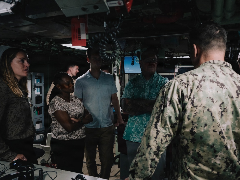 A group of civilians is being briefed or given a tour by a U.S. Navy sailor in digital camouflage (NWU Type III) inside what appears to be a naval vessel's interior compartment or bridge area. The group includes approximately five civilians of mixed gender and ethnicity listening attentively to the uniformed service member. The setting suggests an official visit, media engagement, or distinguished visitor tour aboard a U.S. Navy ship.