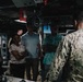 A group of civilians is being briefed or given a tour by a U.S. Navy sailor in digital camouflage (NWU Type III) inside what appears to be a naval vessel's interior compartment or bridge area. The group includes approximately five civilians of mixed gender and ethnicity listening attentively to the uniformed service member. The setting suggests an official visit, media engagement, or distinguished visitor tour aboard a U.S. Navy ship.