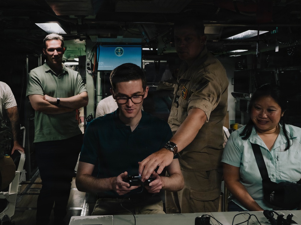 A group of people are gathered in what appears to be a military vessel or facility interior. A young man wearing glasses is seated and holding what appears to be a game controller or remote device, while a person in military/naval uniform leans over to assist or demonstrate. Others observe the activity with smiles.