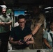 A group of people are gathered in what appears to be a military vessel or facility interior. A young man wearing glasses is seated and holding what appears to be a game controller or remote device, while a person in military/naval uniform leans over to assist or demonstrate. Others observe the activity with smiles.