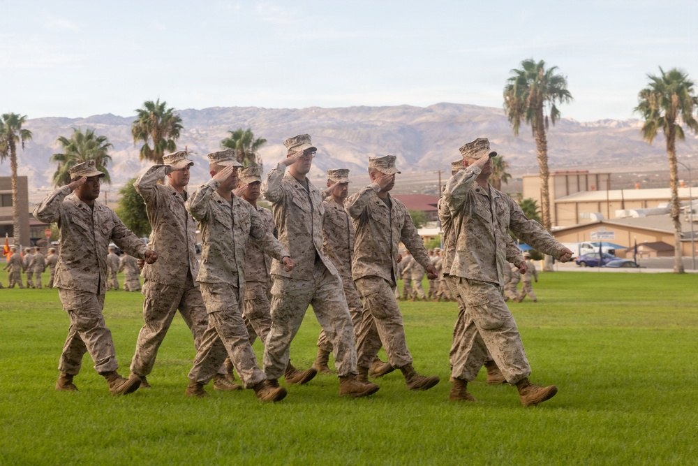 A group of U.S. Marines in desert MARPAT camouflage uniforms are marching in formation across a grass parade field while rendering a salute. The setting appears to be a military installation in a desert region, likely in the American Southwest, identifiable by the arid mountains, palm trees, and base buildings in the background. This appears to be a formal military ceremony or parade review.