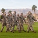 A group of U.S. Marines in desert MARPAT camouflage uniforms are marching in formation across a grass parade field while rendering a salute. The setting appears to be a military installation in a desert region, likely in the American Southwest, identifiable by the arid mountains, palm trees, and base buildings in the background. This appears to be a formal military ceremony or parade review.