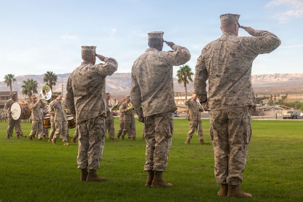 Three U.S. Marines in digital camouflage uniforms (MARPAT) stand at salute on a grass parade field while a military marching band passes in review behind them. The setting appears to be a formal military ceremony, likely a change of command, pass in review, or similar official event. The arid desert landscape and palm trees in the background suggest a southwestern U.S. military installation, consistent with locations such as Twentynine Palms or a similar desert base.