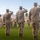 Three U.S. Marines in digital camouflage uniforms (MARPAT) stand at salute on a grass parade field while a military marching band passes in review behind them. The setting appears to be a formal military ceremony, likely a change of command, pass in review, or similar official event. The arid desert landscape and palm trees in the background suggest a southwestern U.S. military installation, consistent with locations such as Twentynine Palms or a similar desert base.