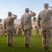 Three U.S. Marines in digital camouflage uniforms (MARPAT) stand at salute on a grass parade field while a military marching band passes in review behind them. The setting appears to be a formal military ceremony, likely a change of command, pass in review, or similar official event. The arid desert landscape and palm trees in the background suggest a southwestern U.S. military installation, consistent with locations such as Twentynine Palms or a similar desert base.