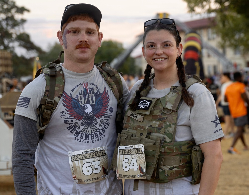 Two participants pose together at a 9/11 Remembrance Run/Ruck/Walk event, wearing tactical plate carriers and race bibs numbered 63 and 64. Both are equipped with rucksacks and wearing military-style gear, with one participant's vest displaying a name tape reading 'SMITH'. The event appears to take place at a military installation, with an inflatable obstacle and crowd visible in the background.