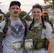 Two participants pose together at a 9/11 Remembrance Run/Ruck/Walk event, wearing tactical plate carriers and race bibs numbered 63 and 64. Both are equipped with rucksacks and wearing military-style gear, with one participant's vest displaying a name tape reading 'SMITH'. The event appears to take place at a military installation, with an inflatable obstacle and crowd visible in the background.