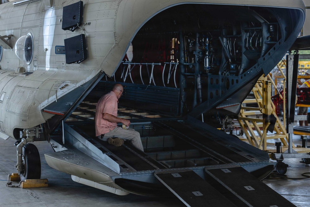 A technician in civilian clothing is seated on the open rear ramp of a large military tandem-rotor helicopter, likely a CH-47 Chinook, inside a maintenance hangar. The aircraft's aft ramp is fully lowered, exposing internal hydraulic lines, structural frames, and mechanical components. The setting appears to be a depot-level or heavy maintenance facility.