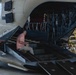 A technician in civilian clothing is seated on the open rear ramp of a large military tandem-rotor helicopter, likely a CH-47 Chinook, inside a maintenance hangar. The aircraft's aft ramp is fully lowered, exposing internal hydraulic lines, structural frames, and mechanical components. The setting appears to be a depot-level or heavy maintenance facility.