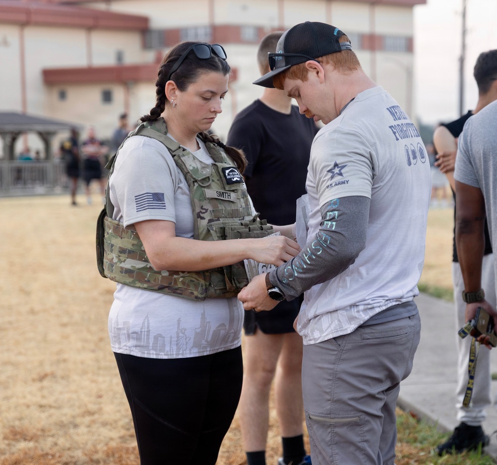 Two individuals are engaged in what appears to be a military or fitness event, with a woman wearing a tactical vest with a name tape reading 'SMITH' interacting with a man in a U.S. Army shirt. The man's shirt reads 'Never Forgotten' and bears U.S. Army branding, suggesting a memorial or commemorative fitness event. Several other participants are visible in the background on what appears to be a military installation.
