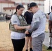 Two individuals are engaged in what appears to be a military or fitness event, with a woman wearing a tactical vest with a name tape reading 'SMITH' interacting with a man in a U.S. Army shirt. The man's shirt reads 'Never Forgotten' and bears U.S. Army branding, suggesting a memorial or commemorative fitness event. Several other participants are visible in the background on what appears to be a military installation.