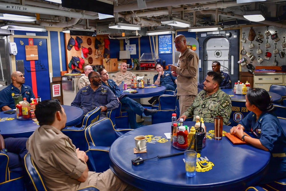 A senior U.S. Navy officer is standing and addressing a group of sailors in what appears to be a Chief Petty Officers' Mess aboard a naval vessel. The room is decorated in blue and gold with Navy insignia, plaques, and unit emblems on the walls. Personnel in various Navy uniforms are seated around round blue tables listening attentively.