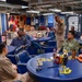 A senior U.S. Navy officer is standing and addressing a group of sailors in what appears to be a Chief Petty Officers' Mess aboard a naval vessel. The room is decorated in blue and gold with Navy insignia, plaques, and unit emblems on the walls. Personnel in various Navy uniforms are seated around round blue tables listening attentively.
