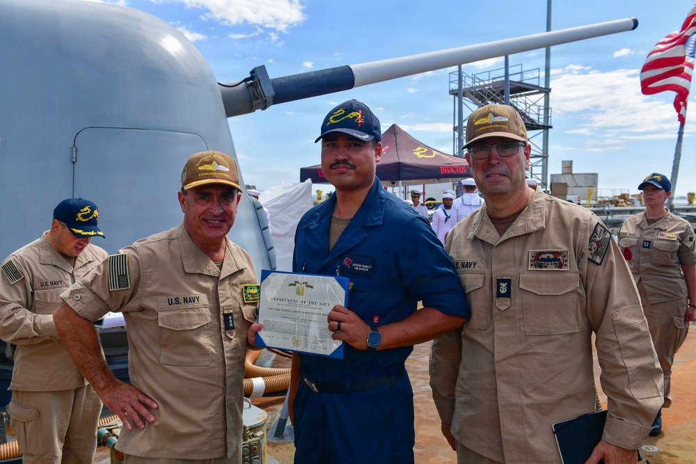 Three U.S. Navy personnel are posing for a photo on the deck of a naval vessel. The central figure in blue Navy working uniform is holding a Department of the Navy certificate, flanked by two senior personnel in tan/khaki uniforms. The setting appears to be an award or recognition ceremony conducted aboard ship.