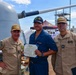 Three U.S. Navy personnel are posing for a photo on the deck of a naval vessel. The central figure in blue Navy working uniform is holding a Department of the Navy certificate, flanked by two senior personnel in tan/khaki uniforms. The setting appears to be an award or recognition ceremony conducted aboard ship.