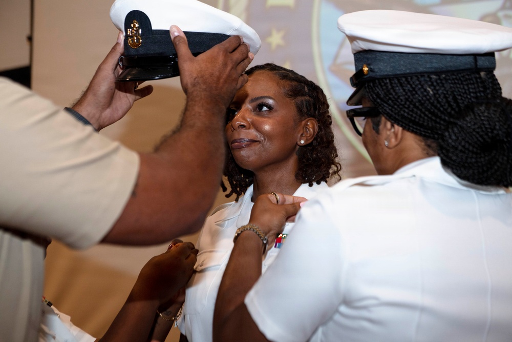 A military promotion or commissioning ceremony is taking place, where a woman in a white uniform is having her cover (hat) placed on her head while another person pins insignia on her collar. The central figure appears emotional, suggesting this is a significant milestone. The setting appears to be an indoor ceremony hall with a projected emblem visible in the background.