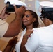 A military promotion or commissioning ceremony is taking place, where a woman in a white uniform is having her cover (hat) placed on her head while another person pins insignia on her collar. The central figure appears emotional, suggesting this is a significant milestone. The setting appears to be an indoor ceremony hall with a projected emblem visible in the background.