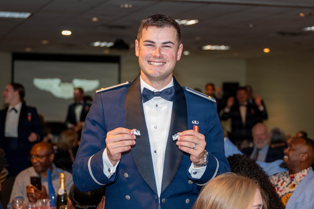 A young man in a military dress uniform (blue formal mess dress with bow tie and epaulettes) is smiling broadly while holding up two small insignia or pins in both hands. The setting appears to be a formal military banquet or dining-in event, with other uniformed personnel and civilians seated at tables in the background. A projection screen is visible in the background, suggesting a formal ceremony or awards presentation.