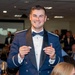 A young man in a military dress uniform (blue formal mess dress with bow tie and epaulettes) is smiling broadly while holding up two small insignia or pins in both hands. The setting appears to be a formal military banquet or dining-in event, with other uniformed personnel and civilians seated at tables in the background. A projection screen is visible in the background, suggesting a formal ceremony or awards presentation.