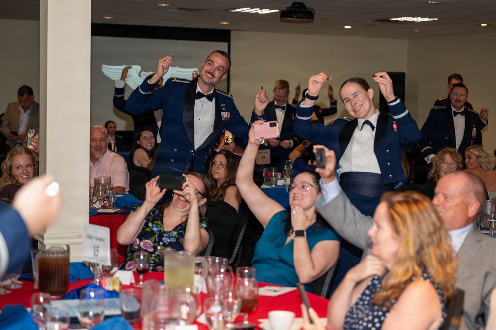A formal military banquet or ball is taking place in a decorated event hall. Two uniformed military personnel in dress blues are standing and celebrating enthusiastically with raised hands, while seated guests photograph the moment. The atmosphere is festive and celebratory, consistent with a military formal dinner or awards ceremony.