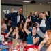 A formal military banquet or ball is taking place in a decorated event hall. Two uniformed military personnel in dress blues are standing and celebrating enthusiastically with raised hands, while seated guests photograph the moment. The atmosphere is festive and celebratory, consistent with a military formal dinner or awards ceremony.