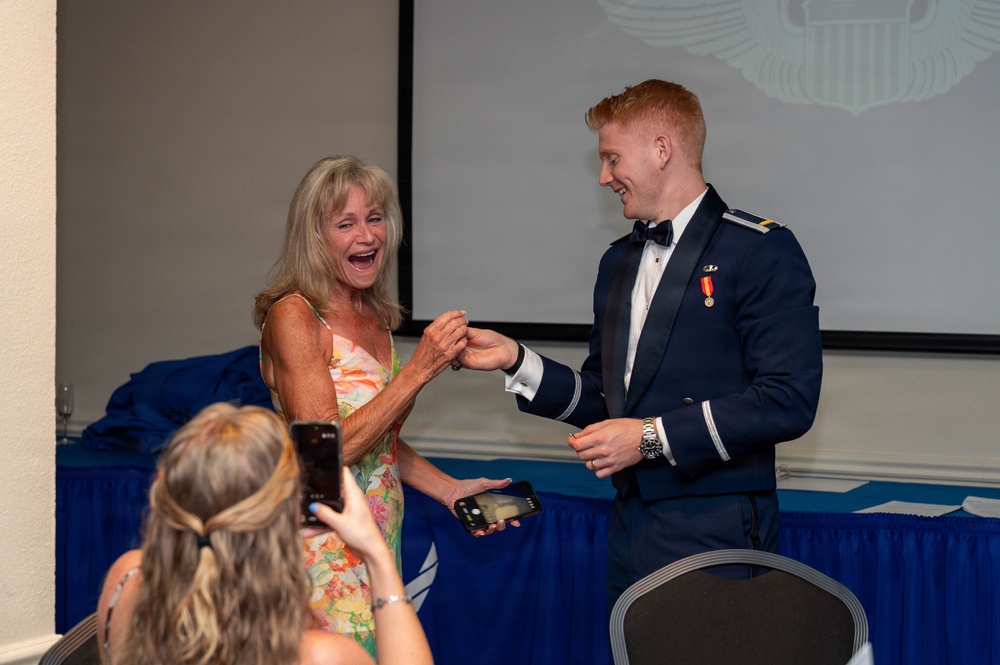 A young man in a military dress uniform is presenting or pinning something to an older woman in a floral dress, who is reacting with visible joy and laughter. The setting appears to be a formal military ceremony or banquet, with a draped table and projection screen visible in the background. A third person in the foreground is capturing the moment on a smartphone.