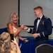 A young man in a military dress uniform is presenting or pinning something to an older woman in a floral dress, who is reacting with visible joy and laughter. The setting appears to be a formal military ceremony or banquet, with a draped table and projection screen visible in the background. A third person in the foreground is capturing the moment on a smartphone.