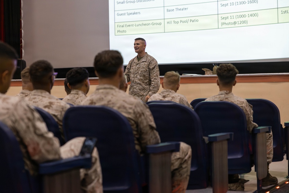 A military briefing or orientation session is taking place in what appears to be a base theater. A senior military officer in MARPAT camouflage uniform stands at the front addressing a seated audience of Marines. A projected schedule slide is visible on the screen behind him, referencing events on September 10-11.