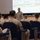 A military briefing or orientation session is taking place in what appears to be a base theater. A senior military officer in MARPAT camouflage uniform stands at the front addressing a seated audience of Marines. A projected schedule slide is visible on the screen behind him, referencing events on September 10-11.