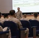 A military briefing or orientation session is taking place in what appears to be a base theater. A senior military officer in MARPAT camouflage uniform stands at the front addressing a seated audience of Marines. A projected schedule slide is visible on the screen behind him, referencing events on September 10-11.